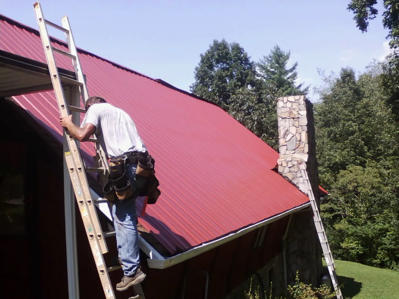 Roofer on a ladder beside a red metal roof and stone chimney — Soffit & Fascia Repair by Roofing Brothers in Crest Hill